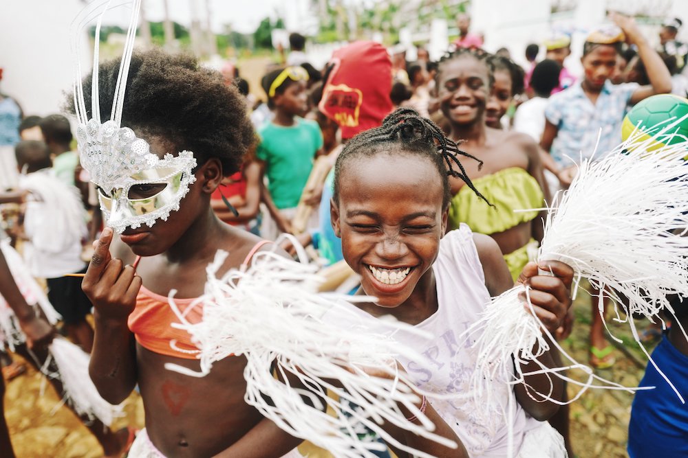 Reise in Sao Tome und Principe. Einheimische Menschen tanzen beim Karneval mit Masken durch die Strassen, im Vordergrund sind glückliche Mädchen zu sehen.